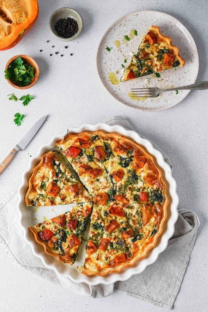 Quiche mit Kürbis, Feta und Spinat auf einem Holztisch serviert.